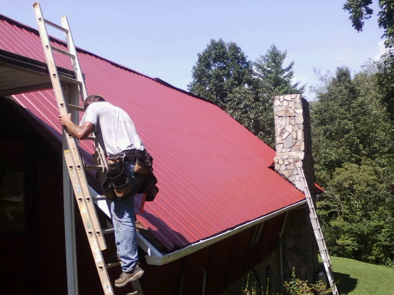 Roofer on a ladder beside a red metal roof and stone chimney — Soffit & Fascia Repair by Roofing Brothers in Aquasco